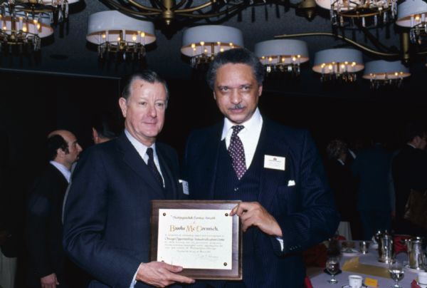 Brooks McCormick standing with Rev. Leon Sullivan. Both men wear International Harvester nametags, and they are holding a plaque which reads, in part: "Distinguished Service Award, Brooks McCormick, In recognition of outstanding support and encouragement to Chicago Opportunities Industrialization Center." Leon Sullivan founded Opportunities Industrialization Centers (OIC) of America. Behind them people are standing around dining tables.