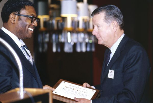 Brooks McCormick receiving an award from a man standing near a podium. The award plaque reads, in part: "Distinguished Service Award, Brooks McCormick, In recognition of outstanding support and encouragement to Chicago Opportunities Industrialization Center." Leon Sullivan founded Opportunities Industrialization Centers (OIC) of America.