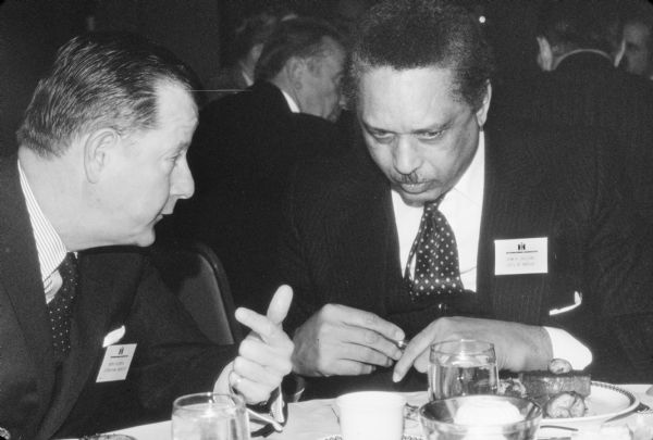 Brooks McCormick sitting and talking with Leon Sullivan at a dinner. The Rev. Leon Sullivan is wearing an International Harvester nametag, with his name and "OIC's of America" on it, which stands for Opportunities Industrialization Centers (OIC) of America. Leon Sullivan founded OIC.