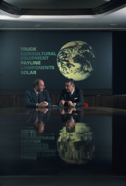 View down long, shiny conference table towards Brooks McCormick and an unidentified man sitting together. On the wall behind them is a sign or projection that reads: "Truck, Agricultural, Equipment, Payline, Components, Solar," along with an image of the earth as seen from space on the right. 