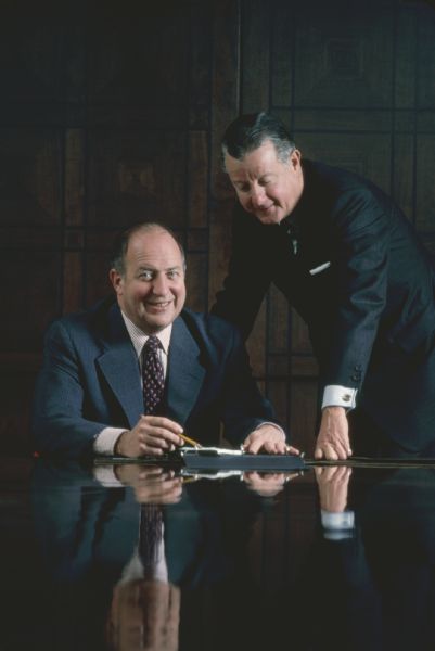 View down long, shiny conference table towards Brooks McCormick and an unidentified man. Brooks is standing and the other man is sitting. The wall behind them is paneled.