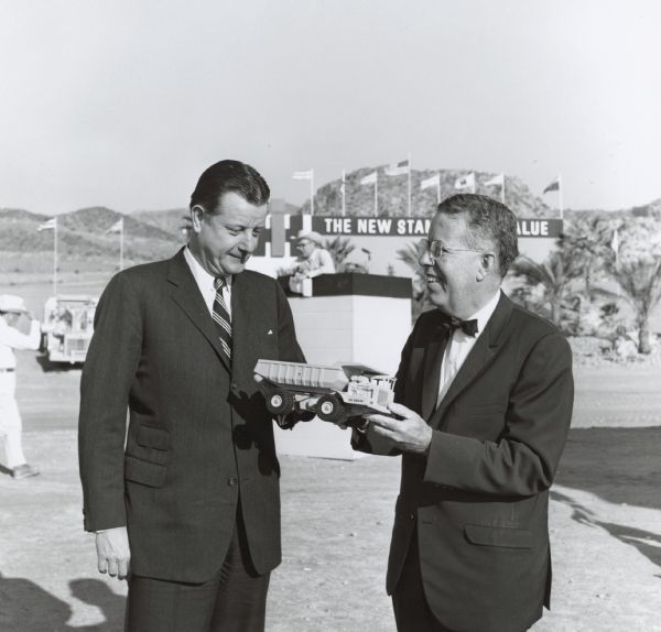 Brooks McCormick standing outdoors next to an unidentified man who is holding an International Pay Hauler toy truck in his hands. Behind them a man stands on a tall box, and in the background is a building with flags, the IH logo, and a sign that reads, in part: "The New St[andard in val]ue." Low hills are in the far background.