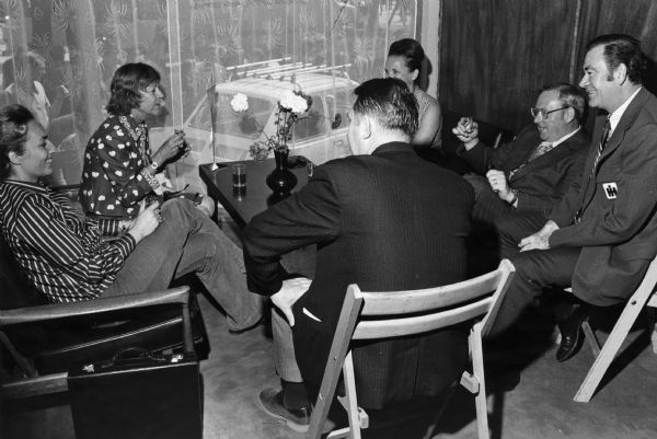 A group of men and women sitting together in a room, relaxing. The man sitting on the far right has an IH name tag on his suit coat. In the background a car is parked just beyond a large window with a curtain. The location may be in Poland.