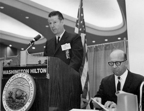 Brooks McCormick stands before a microphone at a podium. The seal of the U.S. Department of the Interior is on the front of the podium, underneath a sign that reads: "Washington Hilton." A man wearing a suit and eyeglasses is sitting on the right.