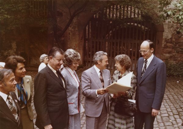 Brooks McCormick stands with a woman on the left, next to a man and woman standing in the center holding open a document together. There is another man standing on the far right, and another man and woman stand on the far left. The group is standing on cobblestones, and behind them is a gated arch in a wall or building.