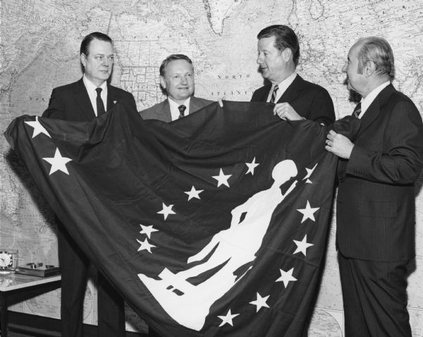 Group portrait of three unidentified men and Brooks McCormick standing and holding a Minuteman flag in front of a large world map.