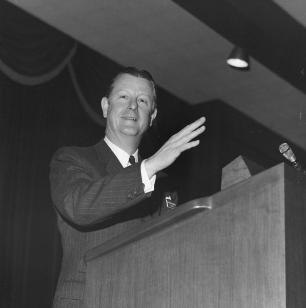 Brooks McCormick standing behind the microphone at a podium with his arm raised in a gesture. The name tag on his suit coat pocket reads: "Brooks McCormick, President."