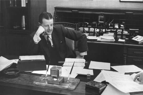 Brooks McCormick sitting behind a desk resting his cheek on his right hand. His elbow is propped on top of the desk blotter. Another person's hands can be seen on top of papers on the desk on the far right. Behind Brooks McCormick is a large, roll-top desk.
