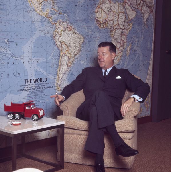 Full-length portrait of Brooks McCormick sitting in a chair holding a gold pen in his right hand. On a table beside him is a red toy dump truck. A large map of the world by "The National Geographic Society" is on the wall behind him.