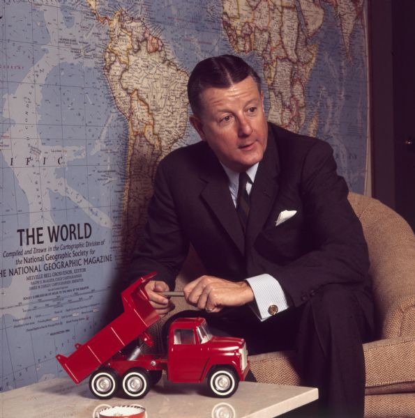 Portrait of Brooks McCormick sitting forward in a chair holding a gold pen in his hands. On the table in front of him is a red toy dump truck. A large map of the world by "The National Geographic Society" is on the wall behind him. 	