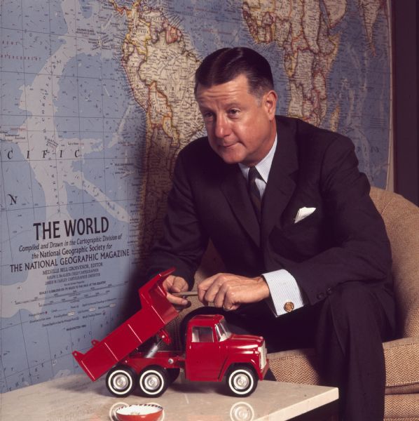 Portrait of Brooks McCormick sitting forward in a chair holding a gold pen in his hands. On the table in front of him is a red toy dump truck. A large map of the world by "The National Geographic Society" is on the wall behind him.