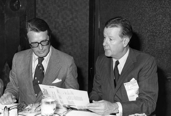 Brooks McCormick sitting at a table at a dinner during the 39th Chicago World Trade Conference. He is sitting next to another man and they are looking at a brochure for the conference. Brooks McCormick is wearing a ribbon on his lapel that reads, in part: "International Harvester Company, Chicago, IL. Chairman."