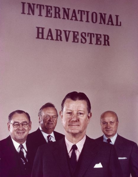 Brooks McCormick is standing in the center foreground with three unidentified men standing behind him. In the background high up on the wall is a sign for "International Harvester."