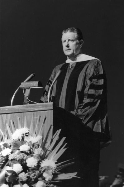 Brooks McCormick standing a podium behind microphones. He is wearing the full-dress gown of someone who has received a honorary degree.
