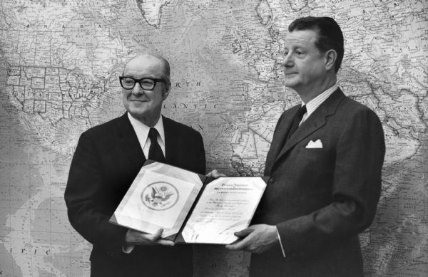 Brooks McCormick, on the right, stands with an unidentified man in front of a large wall map. Together they are holding a folder which has, on the left, the Great Seal of the United States, and on the right, a document with the heading "Treasury Department."