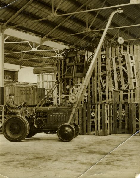 McCormick-Deering tractor equipped with railway wheels and lift or winch attachment driven by a man in a warehouse in New Zealand. In the background is a crate marked "International Speed Truck," and a tall pile of crates hold wheels and other parts.