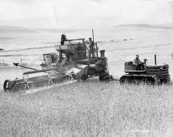 View across field towards a man using a New Zealand International TD-9 TracTractor to pull a man on a McCormick-Deering Harvester Thresher.