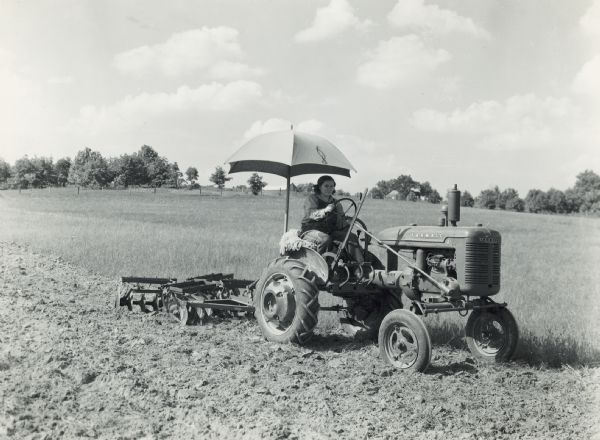 Mrs. Mabel Arbogast of Eldon, Mo., driving a Farmall A pulling a disk harrow through rye on the farm of Mr. E. Arbogast. She is sitting on a sheepskin seat cover under an umbrella attached to the back of the tractor. Farm buildings are in the background.