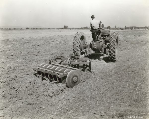 Three-quarter view from right rear of Raymond Ritter of Amity, Oregon, on a Farmall M with a Dyrr off-set disk harrow in 40-acre field covered with straw from a combined heavy vetch seed crop.