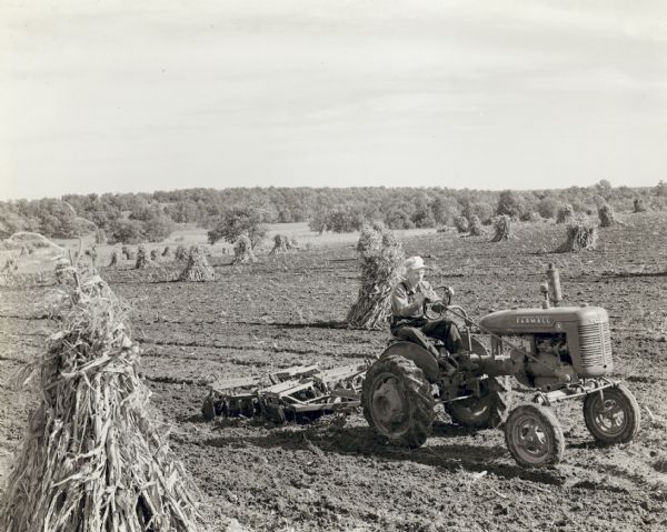 Slightly elevated three-quarter view from right front of a man driving a Farmall A tractor with a disk harrow through a field dotted with corn shocks. In the background is a tree-covered hill. 