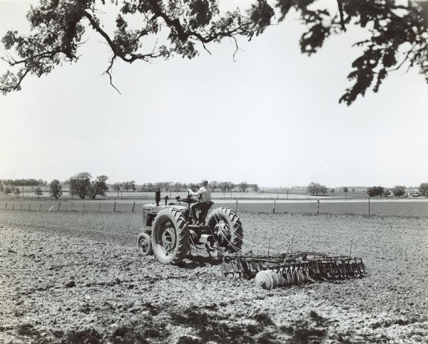Three-quarter view from left rear of a young boy driving a Farmall H which is pulling a disk harrow through a field at the farm of Leslie Winne.