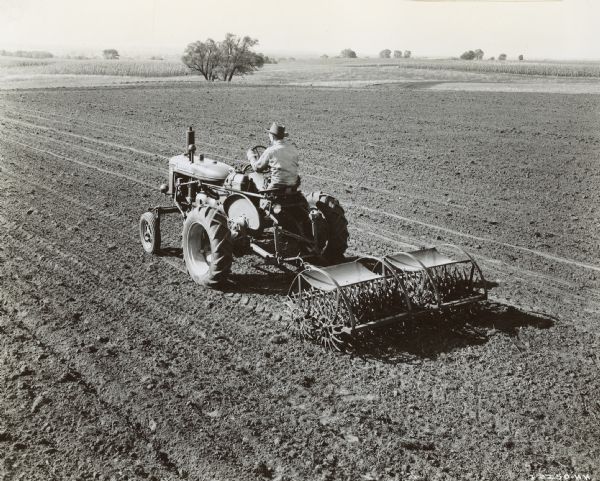 Elevated view from left rear of a man driving an experimental Farmall A pulling a No. 7 rotary hoe in a field.