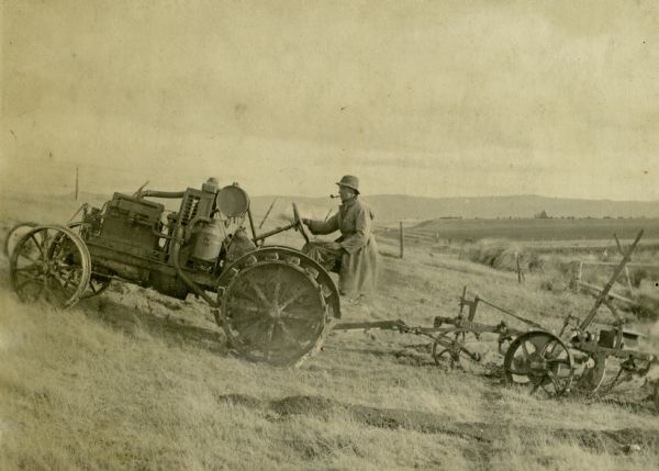 Left side profile view of a man smoking a pipe while driving an International 10-20 tractor, without a engine cover, up a small hill in New Zealand. The tractor is pulling an agricultural implement.