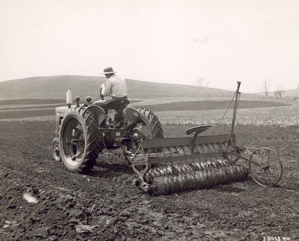 Three-quarter view from left rear of a man driving a Farmall H with soil pulverizers, with grass seeding attachment and seed box. Farm of Tom Stolen, Route 1.