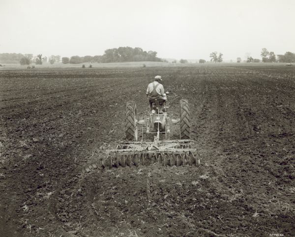 Rear view of man driving a Farmall M tractor with a 9A disk in a field. Owned by Zora Tackett.