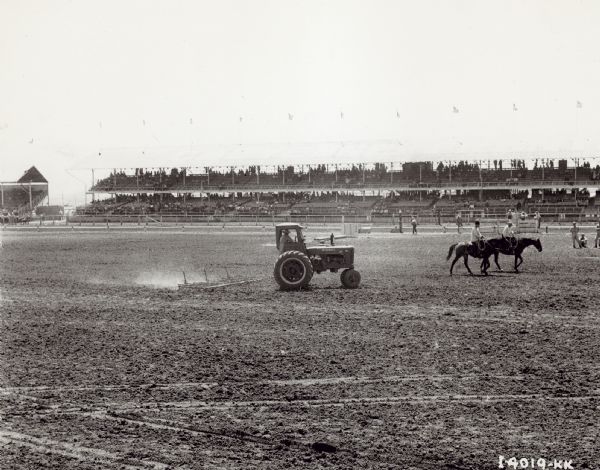 View across ring towards a Farmall M pulling a peg-tooth harrow at the Cheyenne Frontier Days rodeo. Two men are riding on horseback near the tractor. People are watching from stands in the background.