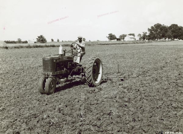 Three-quarter view from left front of a Farmall H tractor with No. 5 peg-tooth harrow preparing ground for oats on the farm of Ed Gentt. Farm buildings are in the background.