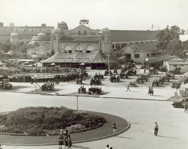 Elevated view of the Texas State Fair, including some of the grounds and a large building with a sign reading: "Automobiles." Tractors are on display in front of the building near a tent and a crowd gathered at a round, fenced-off area with banners that read: "International Harvester" and "International Harvester Speed-Way."