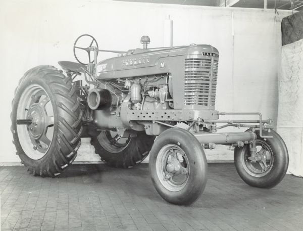 Three-quarter view from right front of a Farmall M tractor with adjustable wide front axle attachment.