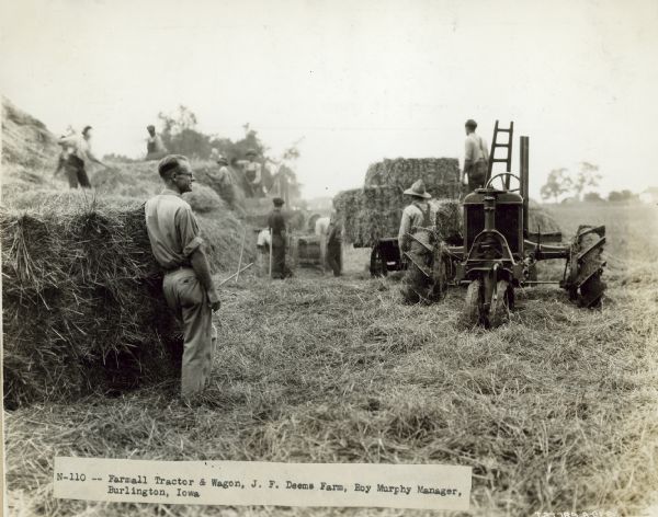 View of men working in a field with a Farmall tractor and wagon. A man stands by a stack of hay bales in the left foreground. Caption: "J.F. Deems Farm, Roy Murphy Manager."