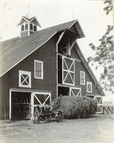 Man driving a Farmall tractor pulling a wagon piled high with hay near the open door of a large barn on Deems Farm. The barn has a cupola with a lightning rod on the roof.