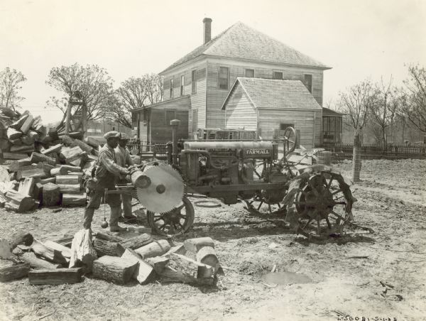 Side view of two men using a Farmall tractor to power a saw that they are using to cut logs. Behind the woodpile is a bell on a small wooden tower. In the background is a farmhouse and farm buildings. A young child is sitting at the top of the back stoop of the house. Oscar E. Wood.