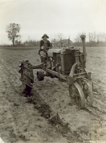 Three-quarter view from front right of a woman driving a Farmall tractor in a field. In the background are farm buildings among trees. W.B. Burkett.