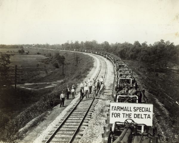 Elevated view of Farmall tractors loaded on railroad cars on railroad tracks next to a group of men standing and posing together on another set of railroad tracks. The locomotive is is in the far background around a bend in the tracks. A sign on the first tractor in the right foreground reads: "Farmall Special for the Delta."