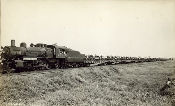 View across field towards the left side of a Chicago Great Western locomotive on railroad tracks pulling cars loaded with Farmall tractors. A sign on the locomotive reads: "International Harvester Tractors." Men are posing looking out of the windows of the locomotive and another man stands at the side of the coal car.