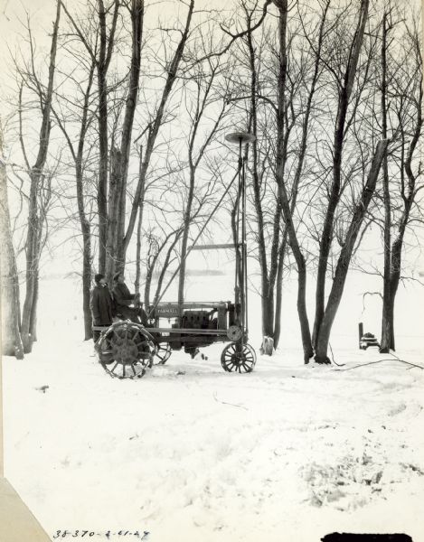 Two men using a Farmall tractor fitted with a circular saw blade mounted high on a tall bracket to trim off the tops of trees. Snow is on the ground. F.G. Beatty.