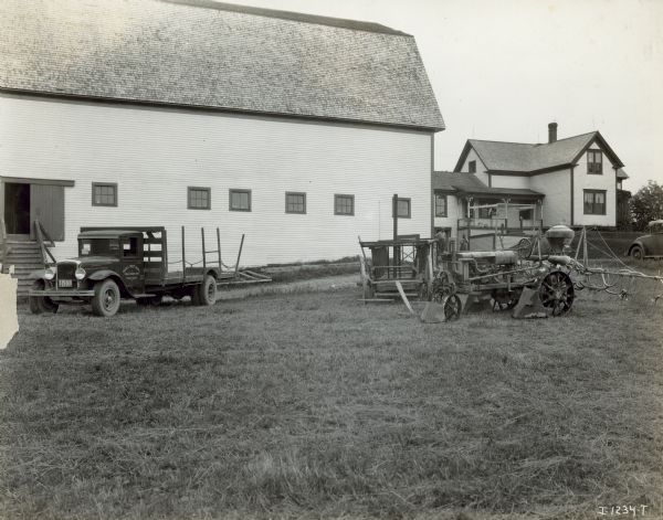 Farmall tractor parked on the farm of C.E. Hussey & Sons. A truck parked on the left has a sign painted on the driver's side door that reads: "Maple Crest Farms, Chas. E. Hussey & Sons." Two men stand in the background between the barn and the farmhouse.