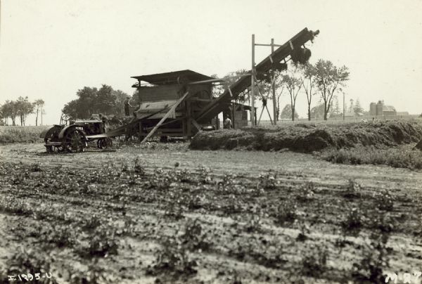 View across field towards men working in a field. A Farmall tractor is belt-driving machinery, perhaps to bale hay. Farm buildings and silos are in the far background. Borg farm. 
