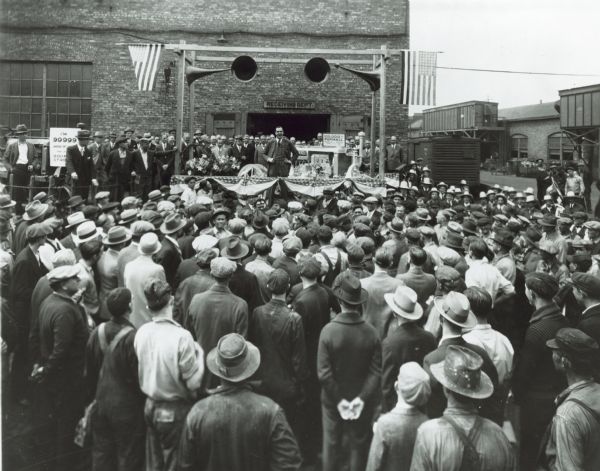 Elevated view over crowd towards an executive who is speaking to workers assembled by the receiving department at Farmall Works to celebrate the 100,000th Farmall tractor. Behind him are the 999,999th and 100,000th tractor.