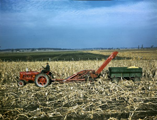 Left side profile view of a man driving a Farmall H tractor in a cornfield with a #14 corn picker.