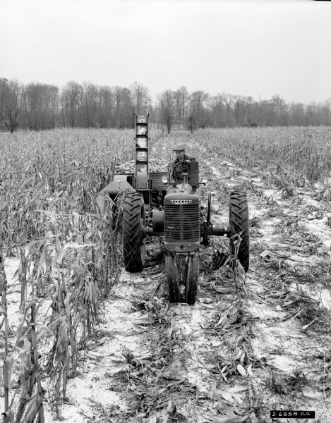 Elevated view from front of a man driving a Farmall H tractor with a 14P corn picker in a cornfield. A light dusting of snow is in the field.