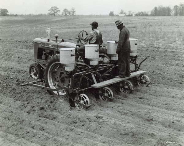 Three-quarter view from left rear of two men using a Farmall M tractor with a four-row cotton planter in a field. Farm buildings are in the far background among trees.