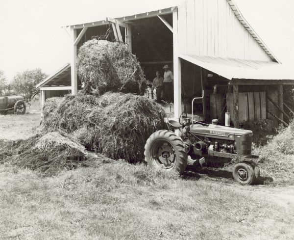 Two men are standing inside an open-sided barn loading hay from a wagon pulled by a Farmall M tractor.