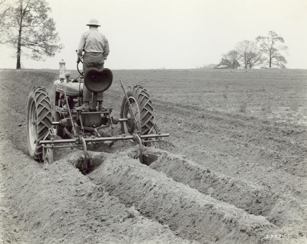 Three-quarter rear view from left of a man standing and driving a Farmall H tractor with an H-9 middle buster in a field. His seat is tipped backwards.