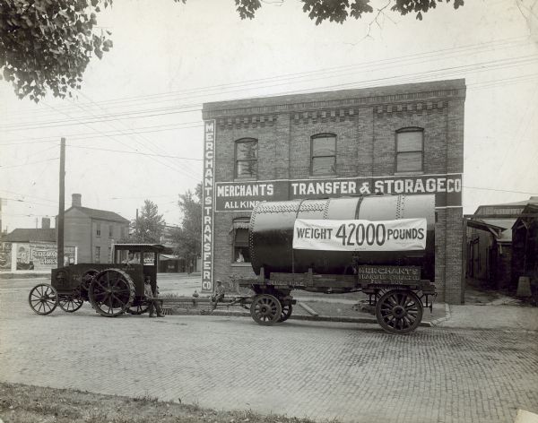 Left side profile view of men with 12-25 Horse Power tractor parked in a cobblestone street. The tractor is pulling a trailer with a large tank that has a banner that reads: "Weight 42000 Pounds." A sign on the trailer reads: "Merchants Transfer & Storage Co., We Move Anything." A young boy is sitting on the long trailer arm in between the tractor and trailer, and a man is looking out of the first floor window of a brick buildng in the background. The sign painted on the building reads: Merchants Transfer & Storage Co." Across the street on the far left are large billboards, with one for "Federal Tires."