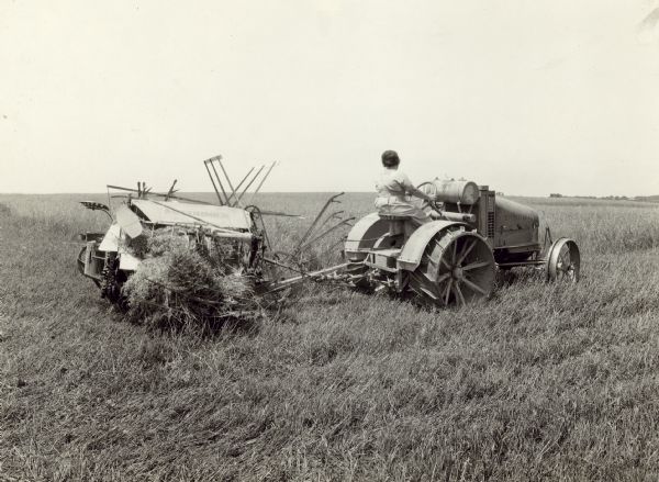 Three-quarter view from right rear of a woman driving a tractor pulling a McCormick harvester thresher in a field. George R. Chester.